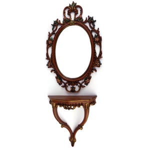 Könnte beinhalten: Ein verzierter, ovaler Spiegel mit passendem Regal. Der Spiegel und das Regal sind braun mit goldenen Akzenten und floralen Details. Der Spiegel ist etwa 51 cm hoch und 30 cm breit. Das Regal ist etwa 25 cm breit.
