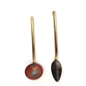 Puede incluir: Dos utensilios de cocina: un cucharón de cobre y una cuchara de metal oscuro, ambos con mangos largos de color latón. El cucharón tiene un cuenco redondo, mientras que la cuchara tiene un cuenco ovalado. Los utensilios están sobre un fondo blanco.