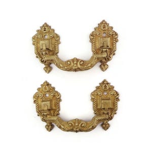 Puede incluir: Dos tiradores dorados ornamentados, de estilo vintage. Cada tirador presenta un asa curvada y detalles decorativos, perfectos para la restauración de muebles antiguos o para añadir un toque de elegancia a cualquier mueble.