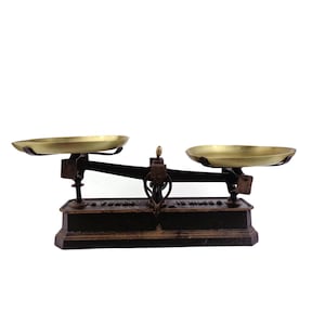 May include: An antique balance scale with two brass pans and a dark metal frame. The scale has a central mechanism with decorative elements and is set against a white background. The scale is a vintage item.