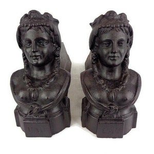 Pode incluir: Um par de suportes de lareira de ferro fundido preto, cada um com um busto de mulher com coroa e tranças. Os suportes têm a forma de um busto de mulher, com a cabeça da mulher voltada para a frente. Os suportes estão em bom estado, sem danos visíveis.