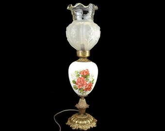 May include: A white lamp with a floral design and a clear glass shade. The lamp has a gold base and a floral design on the shade.