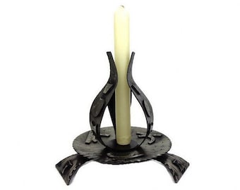 May include: A black metal candle holder with a unique design. The holder has a circular base with three legs and a central stem with two curved arms that hold a white candle.