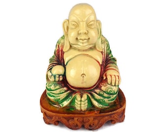 May include: A white ceramic figurine of a smiling Buddha sitting cross-legged on a wooden base. The Buddha is wearing a green and red robe with intricate details.