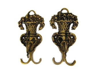 May include: Two antique brass wall hooks. Each hook features a floral basket design at the top, with a decorative body and two hooks at the bottom. The hooks are made of brass and have an aged, vintage appearance.