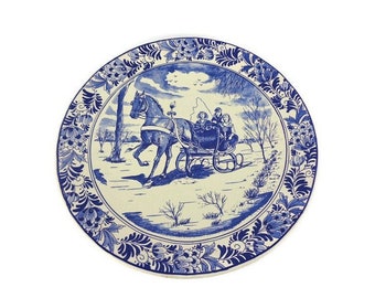 May include: A blue and white ceramic plate with a Dutch Delft design. The plate features a horse-drawn sleigh with three people riding in it. The scene is set in a winter landscape with trees and snow.