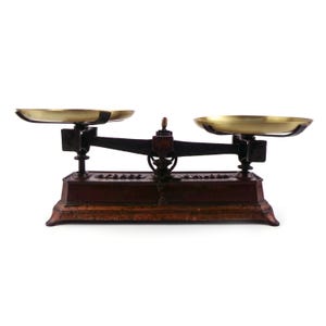 May include: A vintage-style balance scale featuring a dark brown, weathered metal base and a central support beam. Two circular, polished brass pans are mounted on either side of the horizontal arm, which is balanced on a central pivot point.