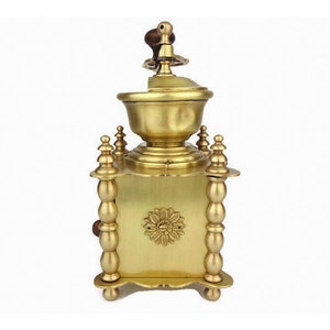 May include: A vintage brass coffee grinder with a wooden handle and ornate details. The grinder has a square base with four legs and a round top with a decorative design.