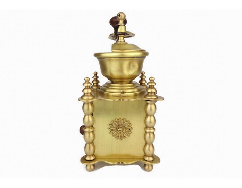May include: A vintage brass coffee grinder with a wooden handle and ornate details. The grinder has a square base with four legs and a round top with a decorative design.
