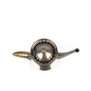 Puede incluir: Una tetera de plata con una tapa decorativa y un asa de madera. La tetera tiene un pico y un cuerpo redondo.
