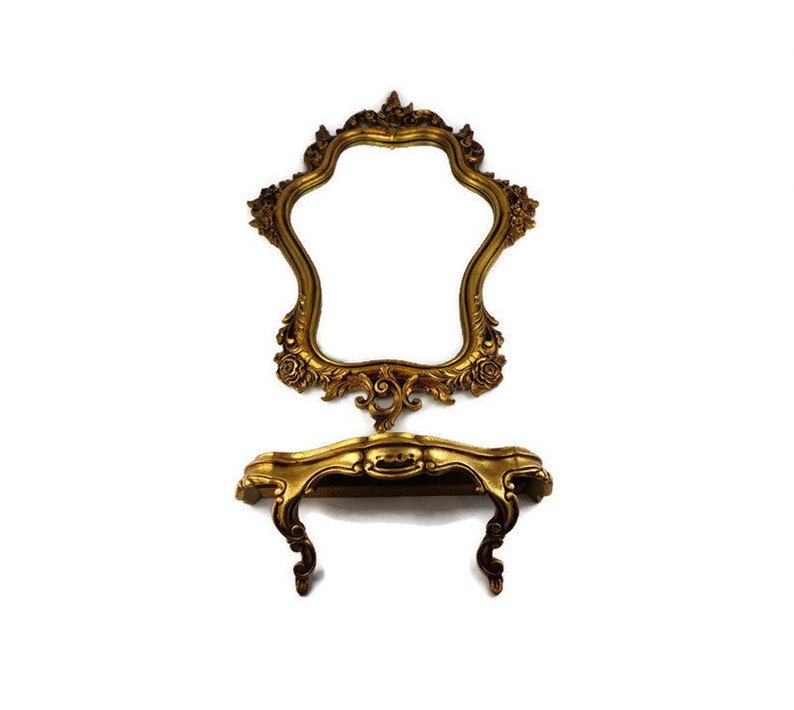 May include: A gold-toned ornate mirror with a decorative shelf underneath. The mirror has a starburst shape and the shelf has a curved design.