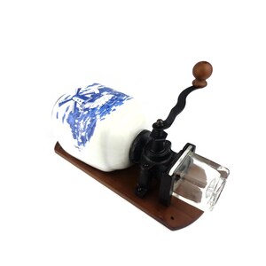 May include: A vintage, wall-mounted coffee grinder with a white ceramic jar featuring a blue and white windmill design. The grinder has a black metal crank and a clear glass hopper.