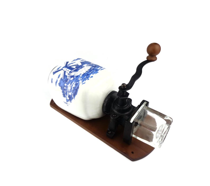 May include: A vintage, wall-mounted coffee grinder with a white ceramic jar featuring a blue and white windmill design. The grinder has a black metal crank and a clear glass hopper.