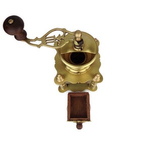 May include: Antique brass coffee grinder with a wooden handle and a drawer for collecting grounds. The grinder has intricate details and a decorative design.