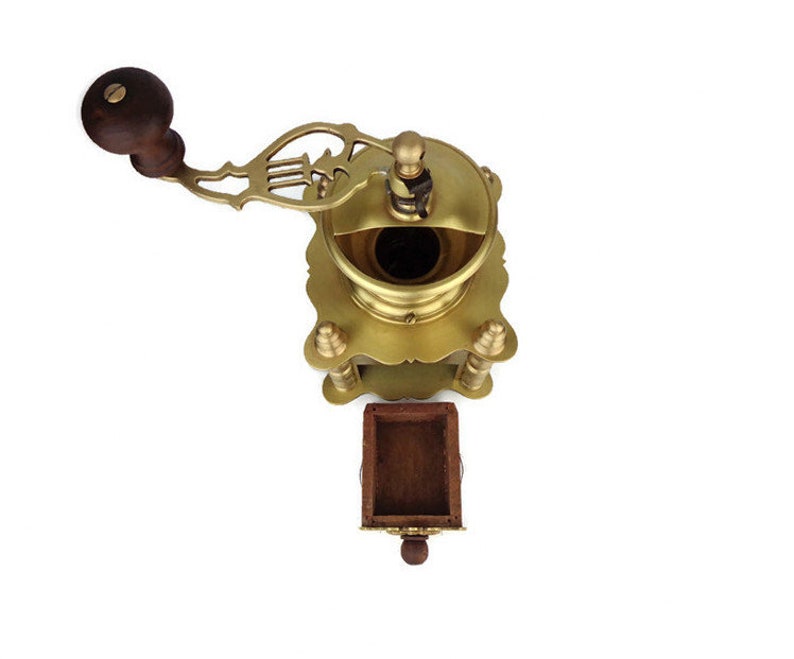 May include: Antique brass coffee grinder with a wooden handle and a drawer for collecting grounds. The grinder has intricate details and a decorative design.