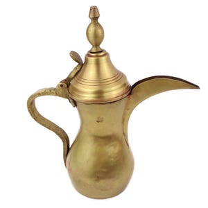 Può includere: Una caffettiera vintage in ottone con beccuccio curvo e manico. La caffettiera ha un coperchio con un pomello decorativo e un'apertura a cerniera. L'ottone ha una calda tonalità dorata, che suggerisce età e utilizzo.