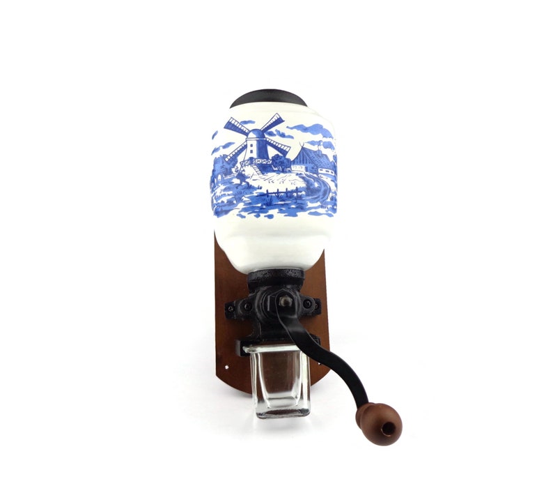 May include: A vintage wall-mounted coffee grinder with a white ceramic jar featuring a blue and white windmill scene. The grinder has a black metal crank and a wooden base.