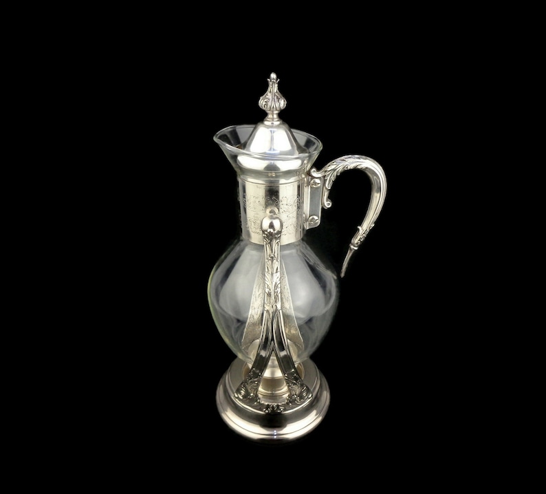 May include: A silver-plated glass pitcher with a decorative handle and a clear glass body. The pitcher has a silver-plated base and a silver-plated lid with a finial.