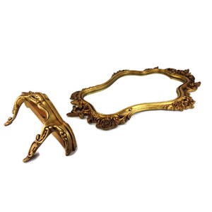 May include: A gold-toned ornate mirror with a decorative stand. The mirror has a wavy, asymmetrical shape and is framed with intricate floral details. The stand is also gold-toned and has a curved, ornate design.