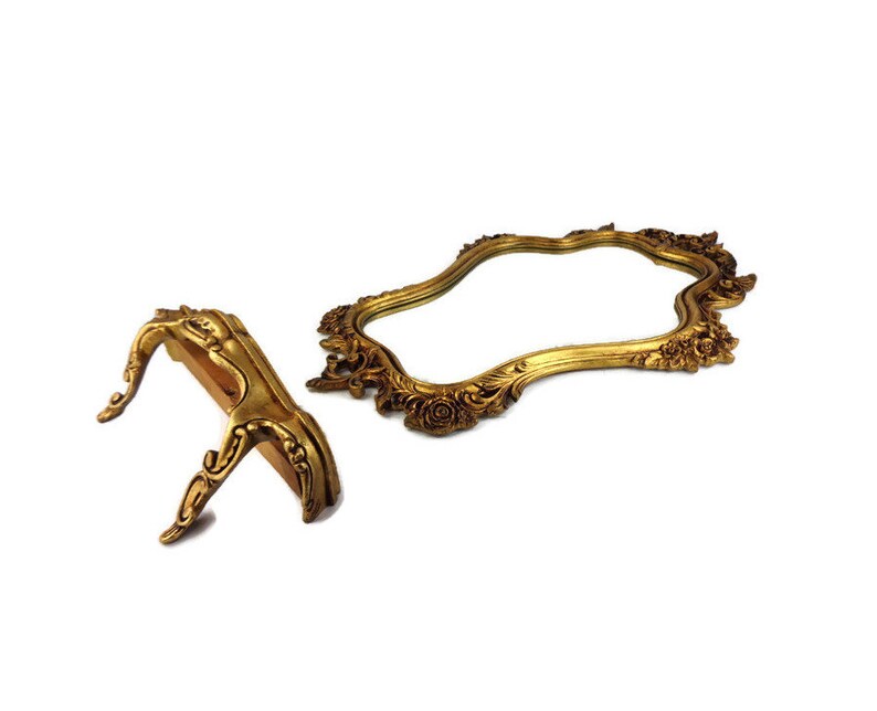 May include: A gold-toned ornate mirror with a decorative stand. The mirror has a wavy, asymmetrical shape and is framed with intricate floral details. The stand is also gold-toned and has a curved, ornate design.