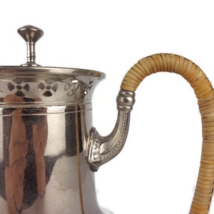 Puede incluir: Una tetera de plata con un asa tejida de color beige. La tetera tiene un dise&ntilde;o decorativo de calado alrededor de la parte superior.