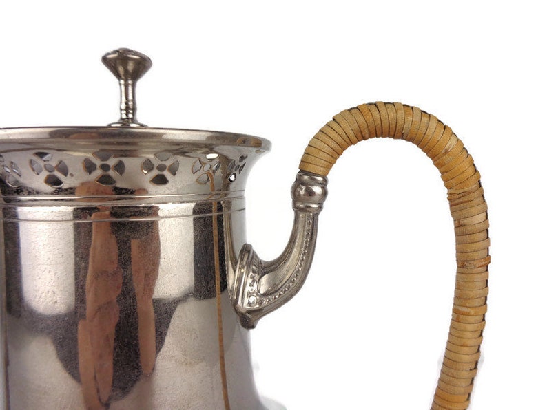 Puede incluir: Una tetera de plata con un asa tejida de color beige. La tetera tiene un dise&ntilde;o decorativo de calado alrededor de la parte superior.