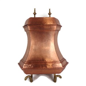 May include: A copper fireplace log holder with a square top and four legs. The holder has a hammered finish and two brass finials on top.