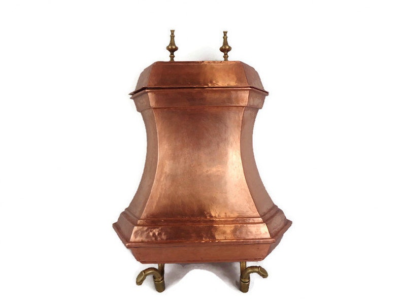 May include: A copper fireplace log holder with a square top and four legs. The holder has a hammered finish and two brass finials on top.