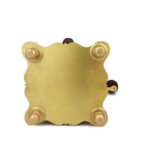 May include: A gold metal stand with four feet and a decorative design. The stand has a round top and a small wooden handle.