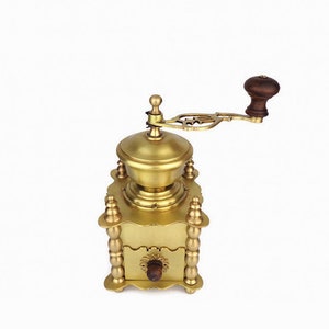 May include: A gold-toned manual coffee grinder with a wooden handle. The grinder has a decorative design and a drawer for collecting coffee grounds.
