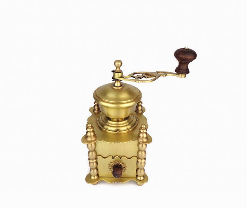 May include: A gold-toned manual coffee grinder with a wooden handle. The grinder has a decorative design and a drawer for collecting coffee grounds.