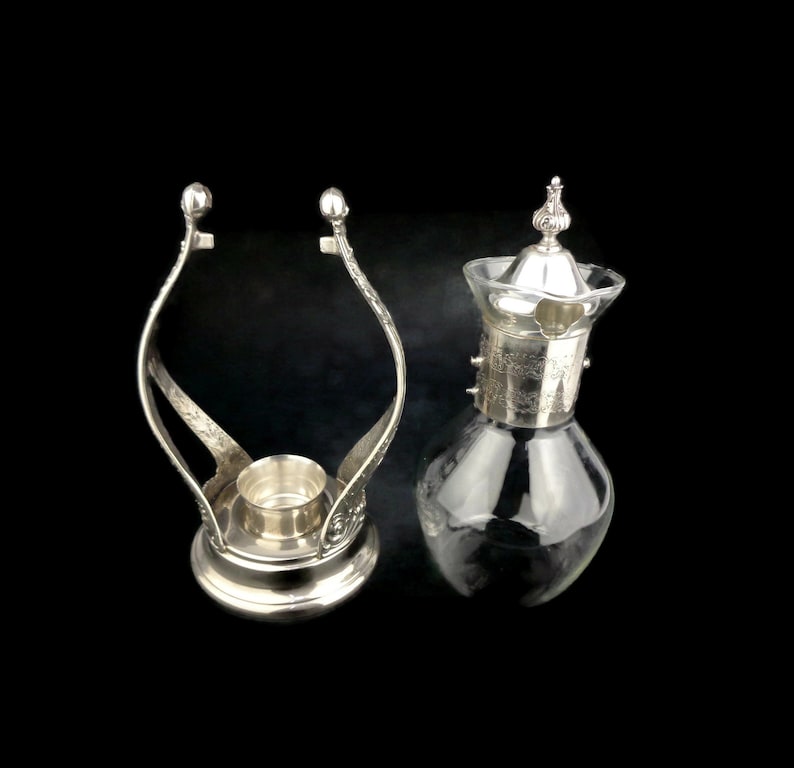 May include: Silver plated candle holder with a glass carafe with a silver lid. The candle holder has a decorative design and the carafe has a floral design.