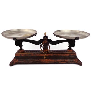 May include: Antique cast iron balance scale with two round metal weighing pans. The scale is painted in shades of red and brown with a worn, distressed finish.