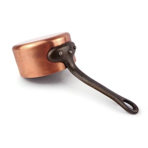 May include: A copper saucepan with a smooth, polished finish and a rounded body. The pan features a dark, textured cast iron handle securely attached to the side with three visible metal rivets, and a circular hole at the end of the handle.
