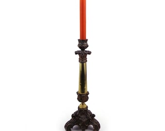 May include: An ornate, antique-style candlestick holder with a tall, orange candle. The base and decorative elements are dark brown, while the central column is gold-colored. The candle is approximately 8 inches tall.