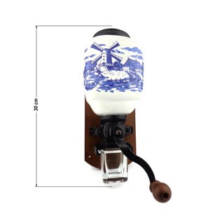 May include: A vintage, wall-mounted coffee grinder with a white ceramic jar featuring a blue and white windmill scene. The grinder has a wooden base and a black metal crank handle. The jar is 30 cm tall.