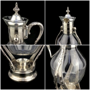 May include: Silver plated glass coffee pot with a decorative stand. The pot has a clear glass body and a silver plated lid with an ornate design. The stand has a round base and three legs.