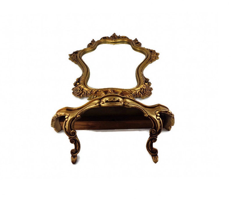 May include: A gold-toned ornate mirror with a shelf underneath. The mirror has a scalloped edge and intricate floral details. The shelf is also decorated with ornate details.