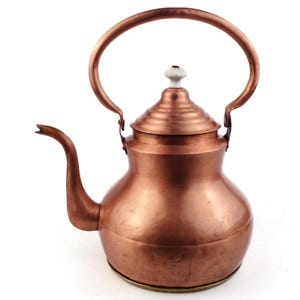 May include: A copper kettle with a large handle and a curved spout. The kettle has a lid with a white knob. The copper has a warm, reddish-brown colour and a shiny finish. The kettle is likely vintage or antique.
