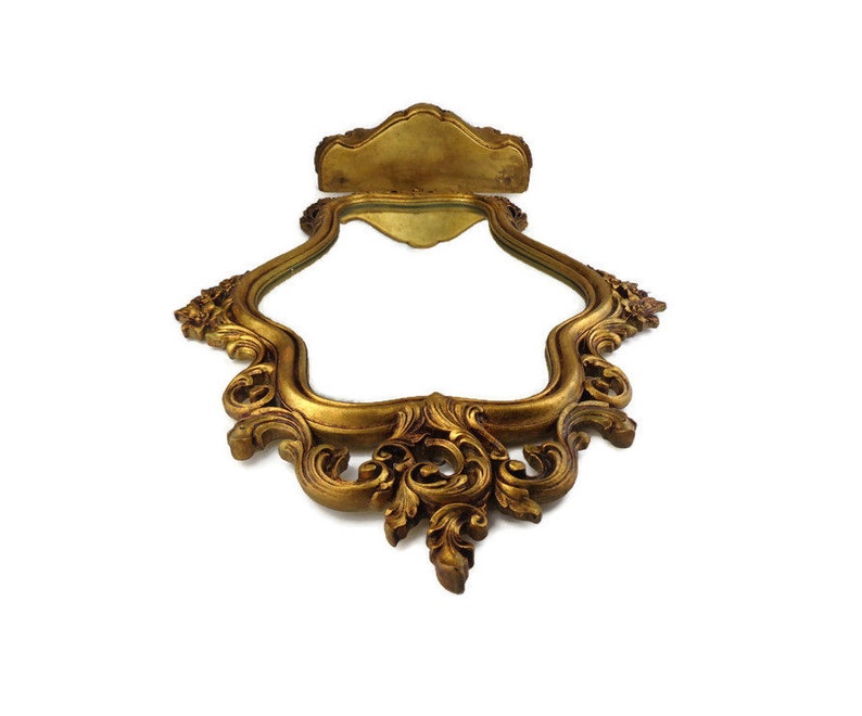 May include: A gold-toned ornate mirror with a decorative frame. The frame has a scalloped edge and intricate floral designs.