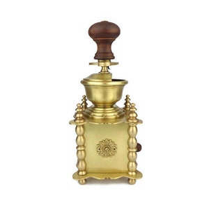 May include: A gold-toned manual coffee grinder with a wooden handle. The grinder has a square base with decorative details and a round top.