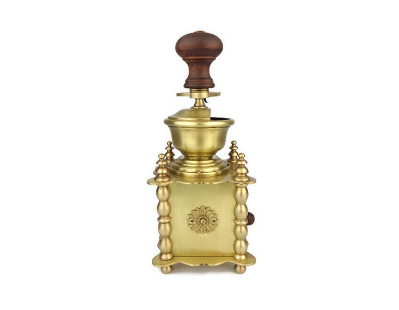 May include: A gold-toned manual coffee grinder with a wooden handle. The grinder has a square base with decorative details and a round top.