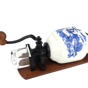 May include: A vintage-style, wall-mounted coffee grinder with a white ceramic jar decorated with a blue and white windmill scene. The grinder has a wooden base and a black metal crank.