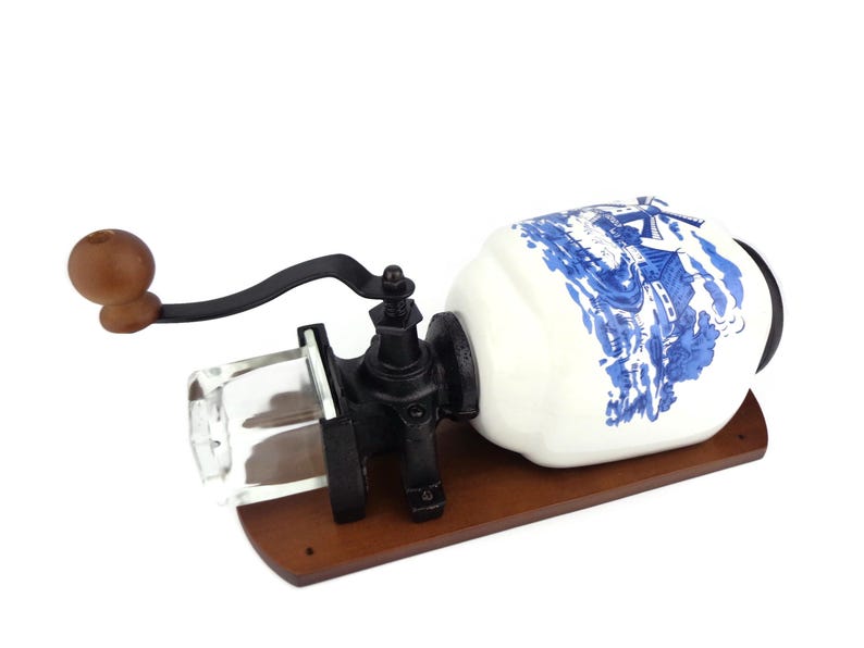May include: A vintage-style, wall-mounted coffee grinder with a white ceramic jar decorated with a blue and white windmill scene. The grinder has a wooden base and a black metal crank.