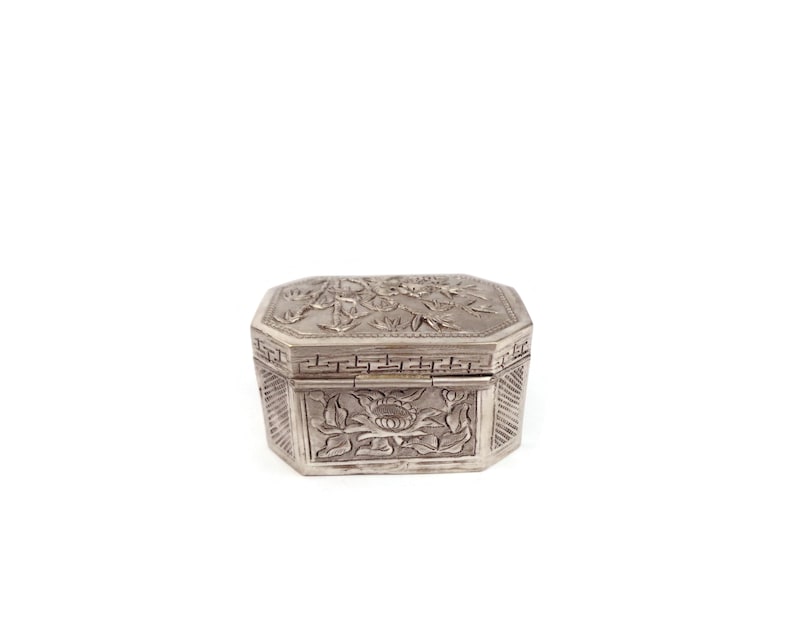 May include: A silver box with an intricate floral design. The box has a hinged lid and is decorated with a repeating pattern of flowers and leaves.