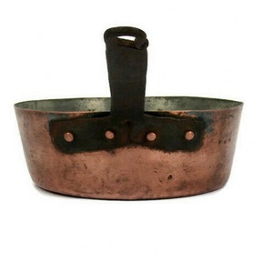 May include: A vintage copper pot with a black iron handle. The pot has a rounded bottom and a wide, shallow opening. The handle is attached to the pot with rivets.