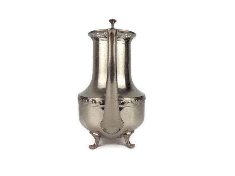 Puede incluir: Una tetera de plata con un dise&ntilde;o decorativo y tres patas. La tetera tiene un pico y una tapa.