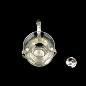 May include: A silver and glass sugar dispenser with a decorative handle and a separate lid. The dispenser is on a black background.