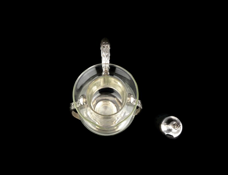 May include: A silver and glass sugar dispenser with a decorative handle and a separate lid. The dispenser is on a black background.