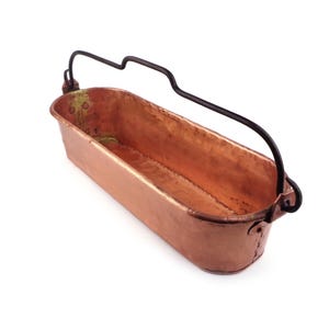 May include: An elongated rectangular copper container with a hammered finish and a dark metal swing handle attached to the sides. The interior shows a warm metallic sheen, while the exterior features visible rivets securing the handle mounts.